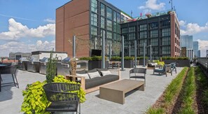 A rooftop patio with outdoor seating, a barbecue grill, and a view of a modern building.