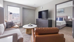 Quarto de hotel moderno com sofá, mesa de centro redonda, poltrona de couro, televisão, cama e mesa de cabeceira.