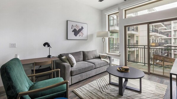 Un salon moderne avec un canapé gris, un fauteuil vert, une petite table ronde et une grande fenêtre donnant sur la façade de l’immeuble.