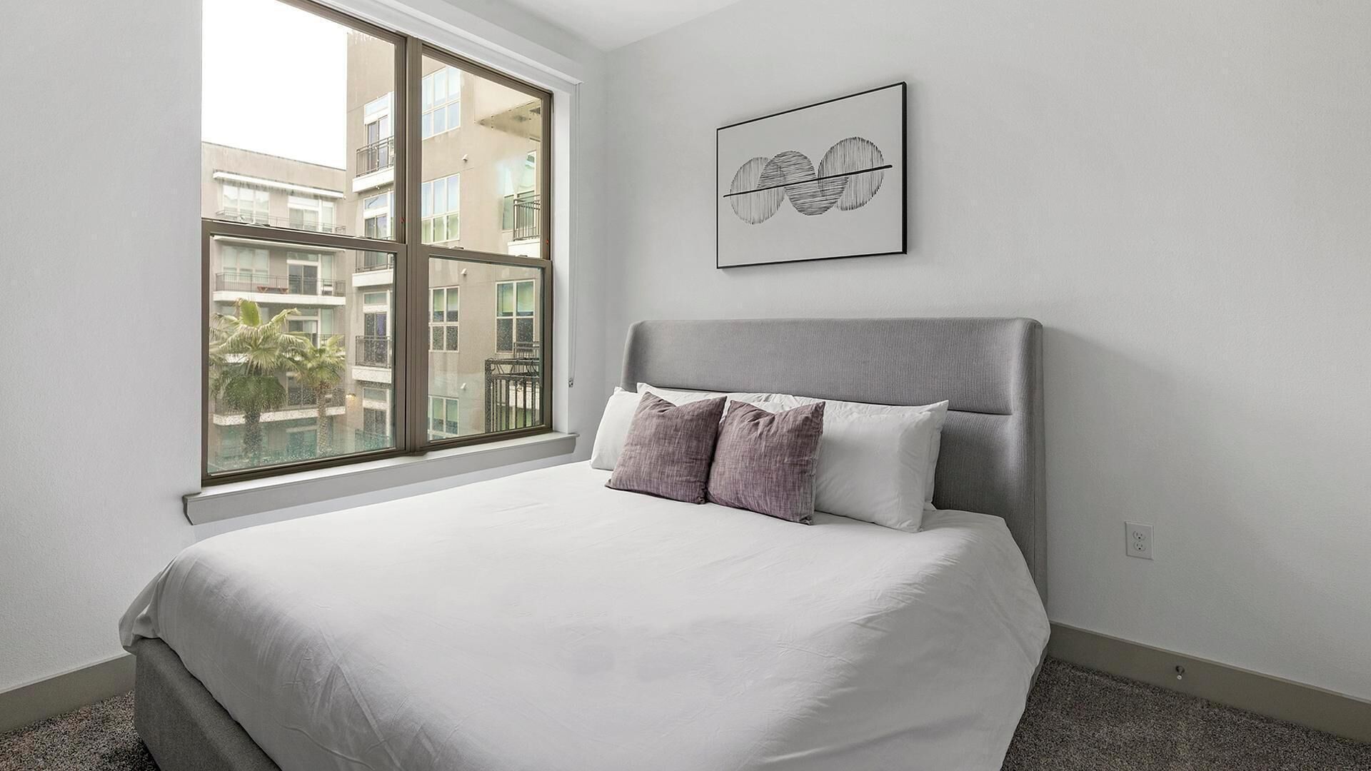 A bedroom with a large window, a bed with white and purple bedding, and a framed artwork on the wall.