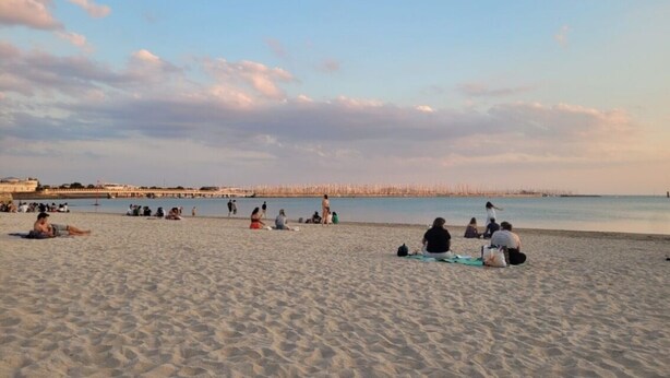 Una playa con gente relajándose en la arena, un mar tranquilo y una costa lejana con edificios.