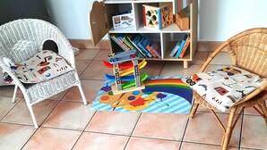 Una habitación con techo inclinado, una estantería, dos sillones de mimbre, un tapete de colores y una variedad de juguetes y libros infantiles.