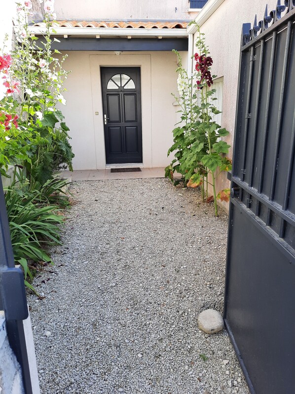 Un camino de grava que conduce a una puerta negra, con una pared blanca y un tejado de baldosas.