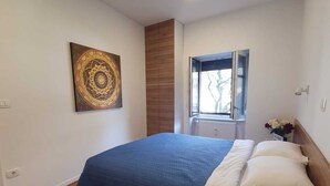 A bedroom with a bed, a wooden wardrobe, a window with blinds, a wall-mounted light, and a decorative wall piece. - Cozy 1-bedroom apartment with WiFi, AC in enjoyable Ljubljana (Ljubljana)