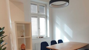 A modern dining area with a wooden table and blue chairs, a white shelving unit, and a large window with blinds. - Cozy 1-bedroom apartment with WiFi, AC in enjoyable Ljubljana (Ljubljana)