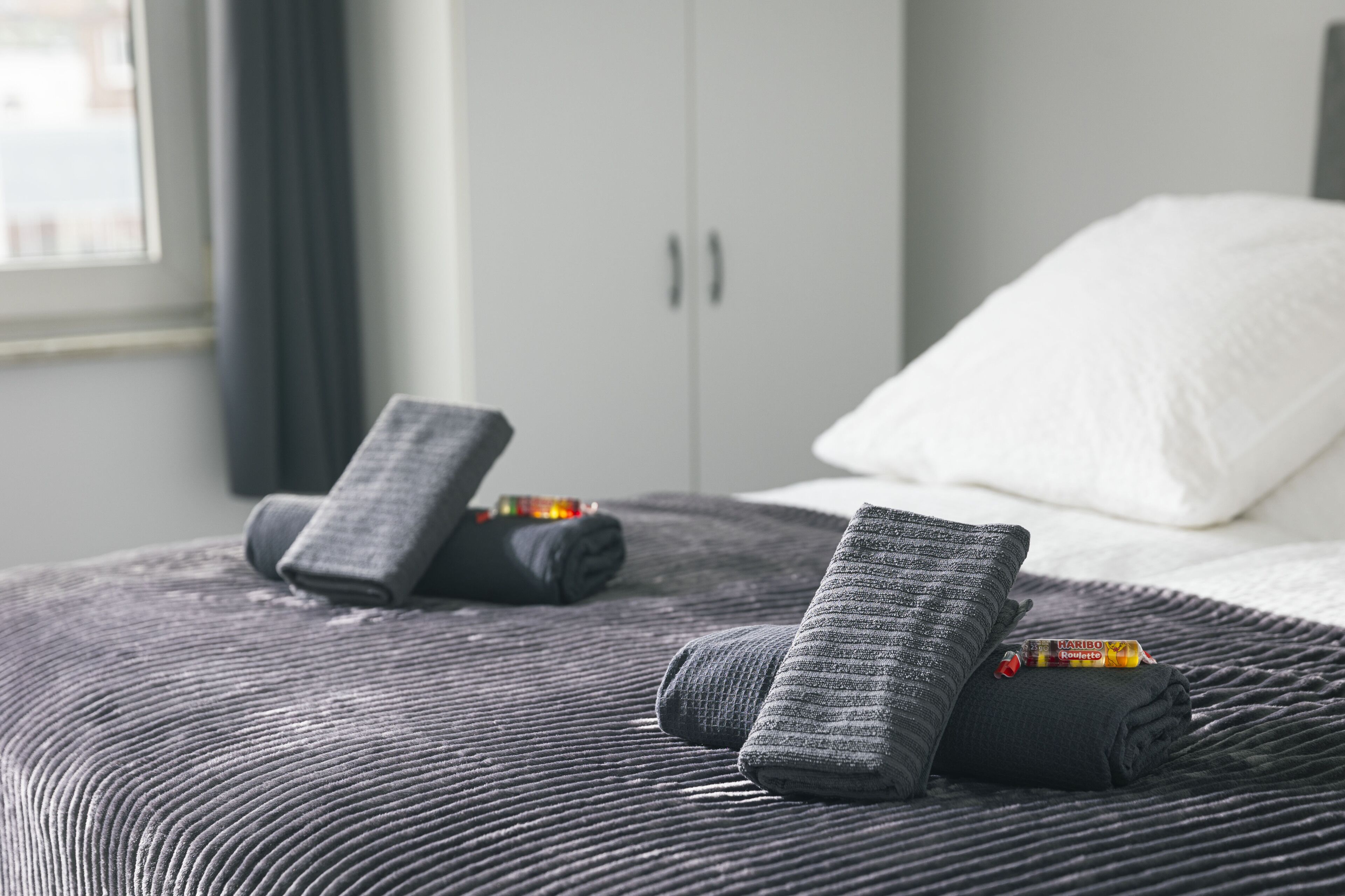 A neatly made bed with a dark textured bedspread, two rolled-up towels, and a small packet of Karibo lotion on the bed.