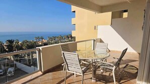 Un balcón con barandilla de vidrio, una mesa con sillas y vista al mar.