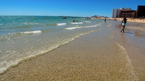 Una playa con agua cristalina, gente caminando y edificios al fondo.