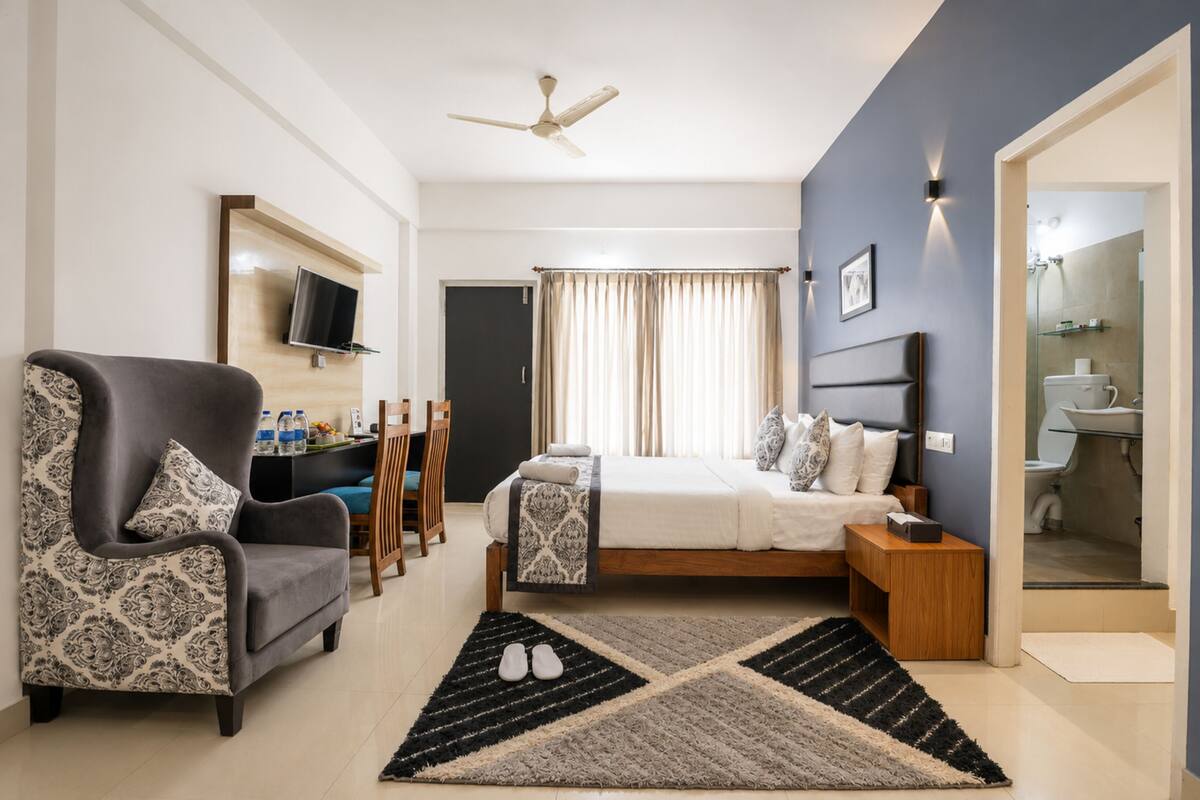Une chambre à coucher avec un lit, une chaise, un téléviseur, un ventilateur de plafond et une salle de bain visible à travers une porte ouverte.