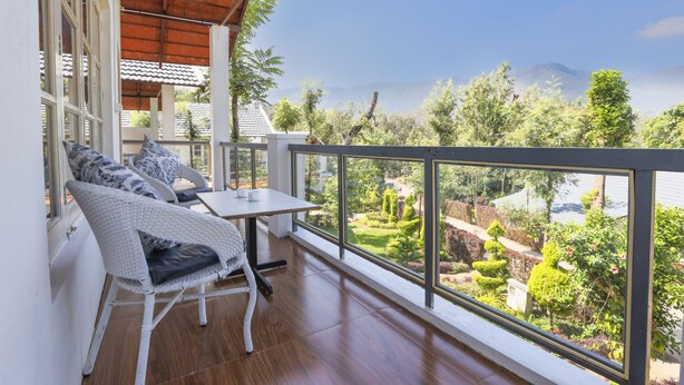 A balcony with wicker furniture and a table, overlooking a well-maintained garden and distant mountains.