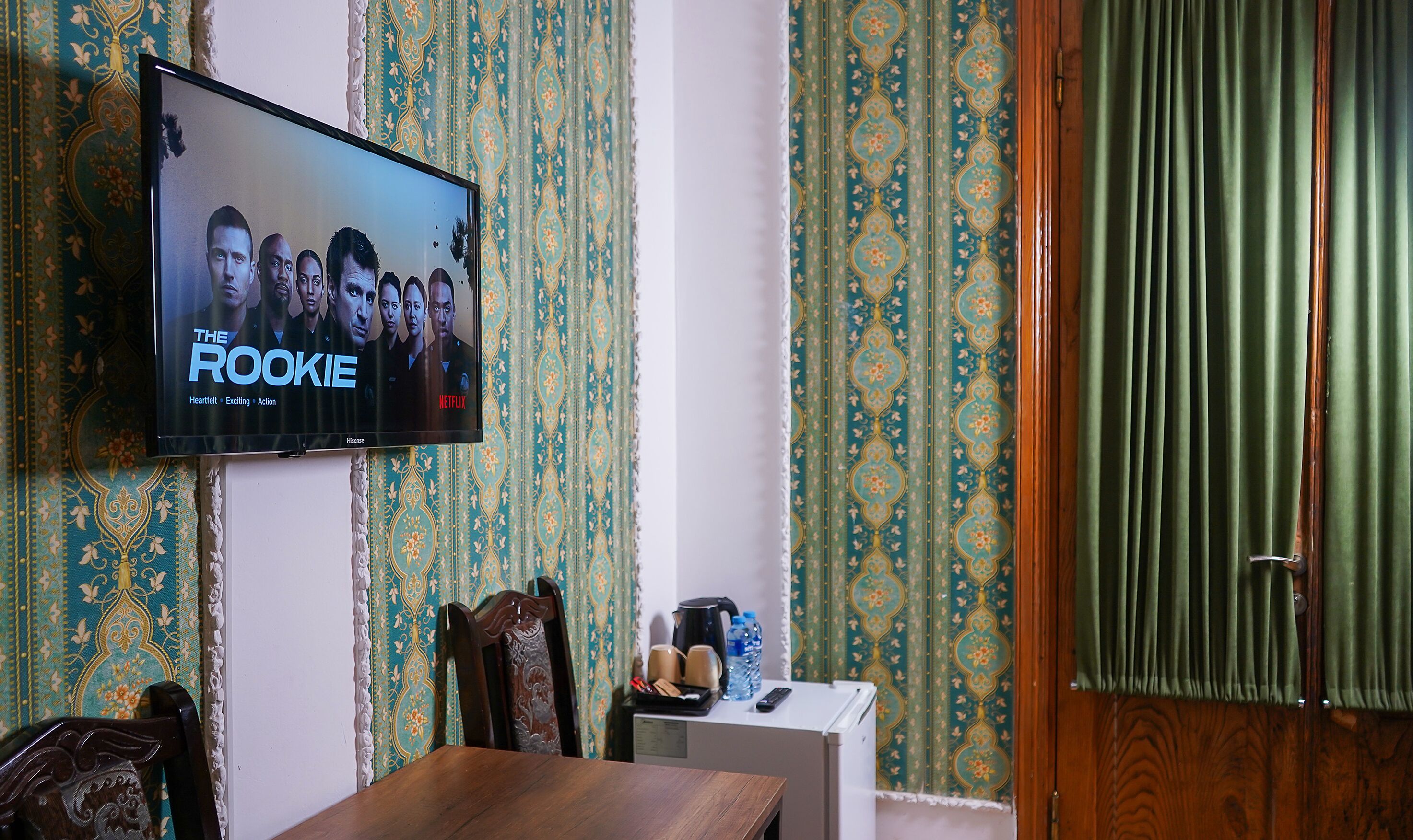 Una habitación con una televisión que muestra 'The Rookie' en Netflix, una mesa de comedor de madera, un armario blanco pequeño y un papel tapiz estampado.