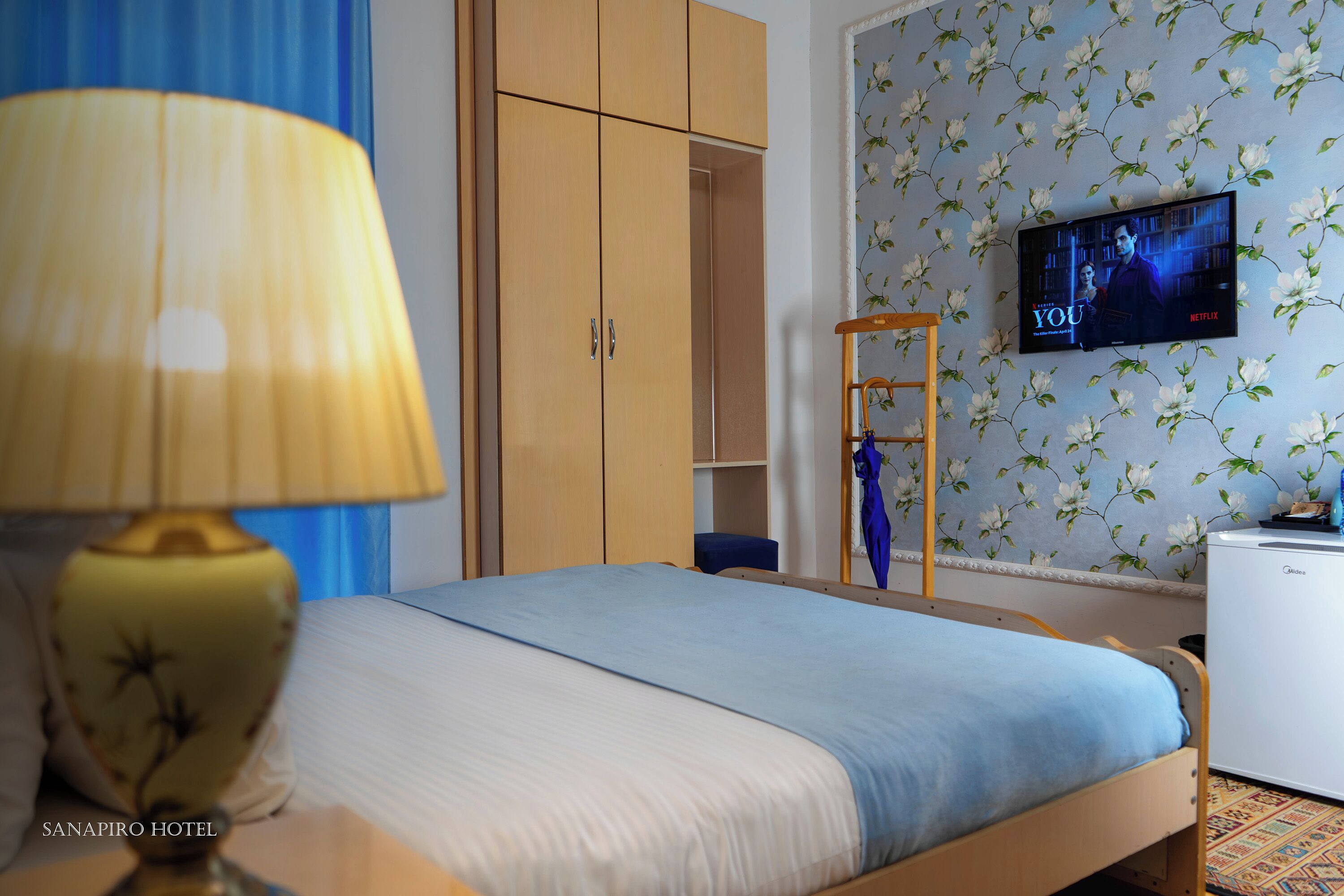 A hotel room with a bed, a TV mounted on the wall, a wardrobe, and a desk with a lamp.