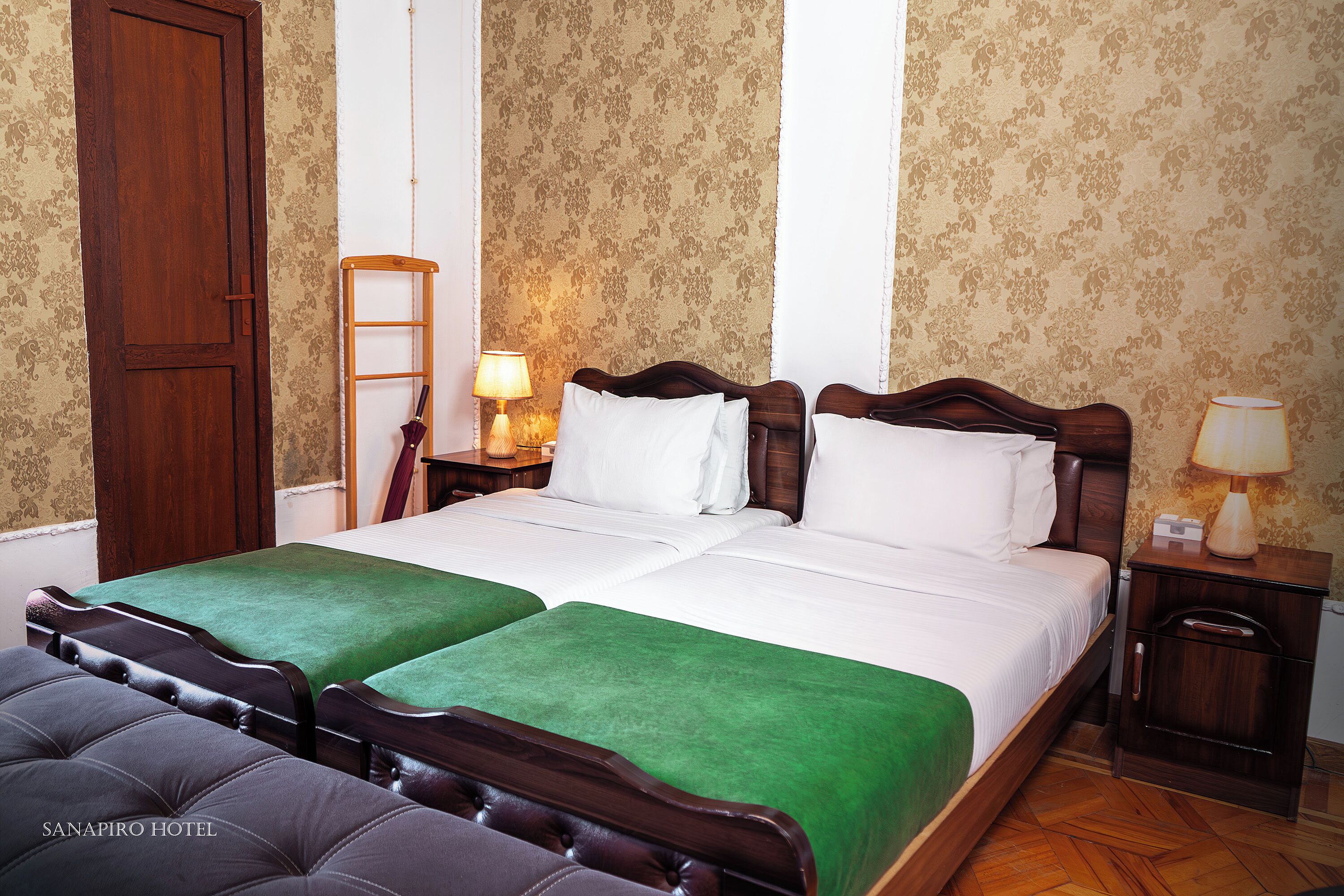 A hotel room with two beds, green bedspreads, wooden headboards, a wooden wardrobe, and a nightstand with a lamp.
