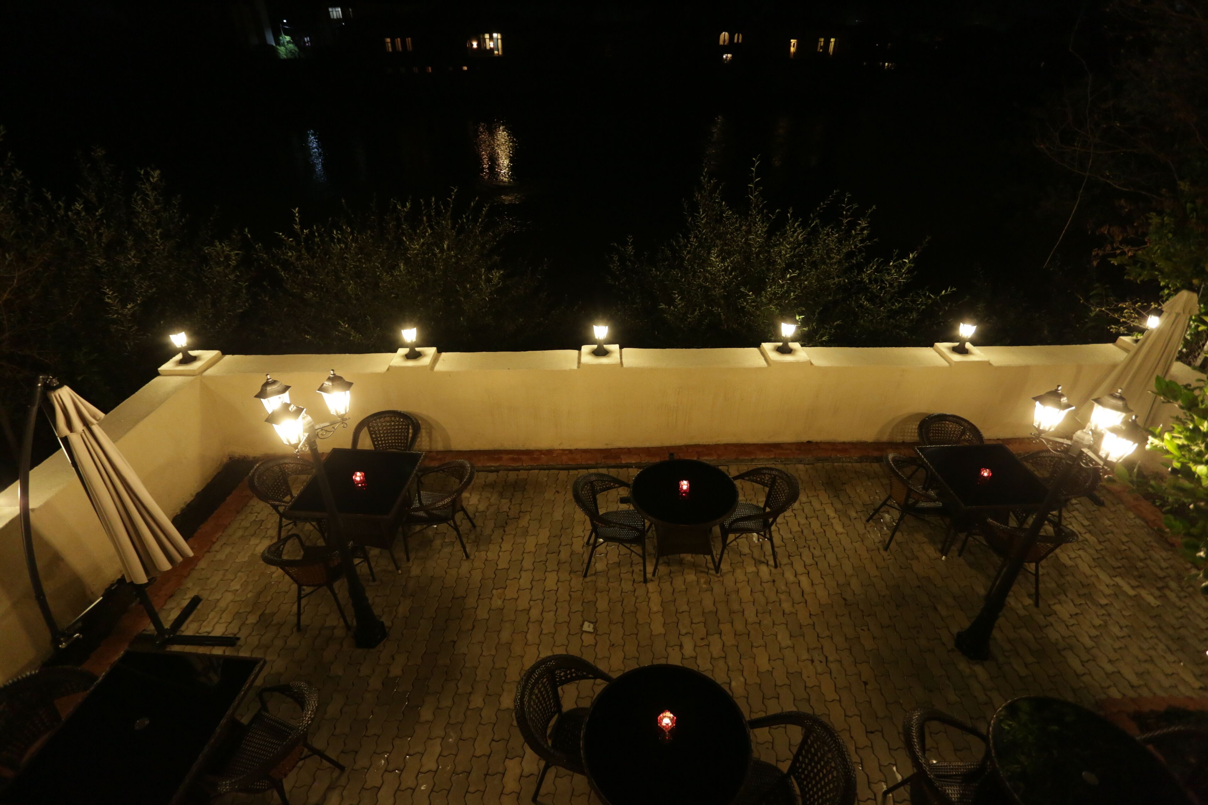 An outdoor patio area with tables and chairs, surrounded by a low wall and a view of a body of water at night.