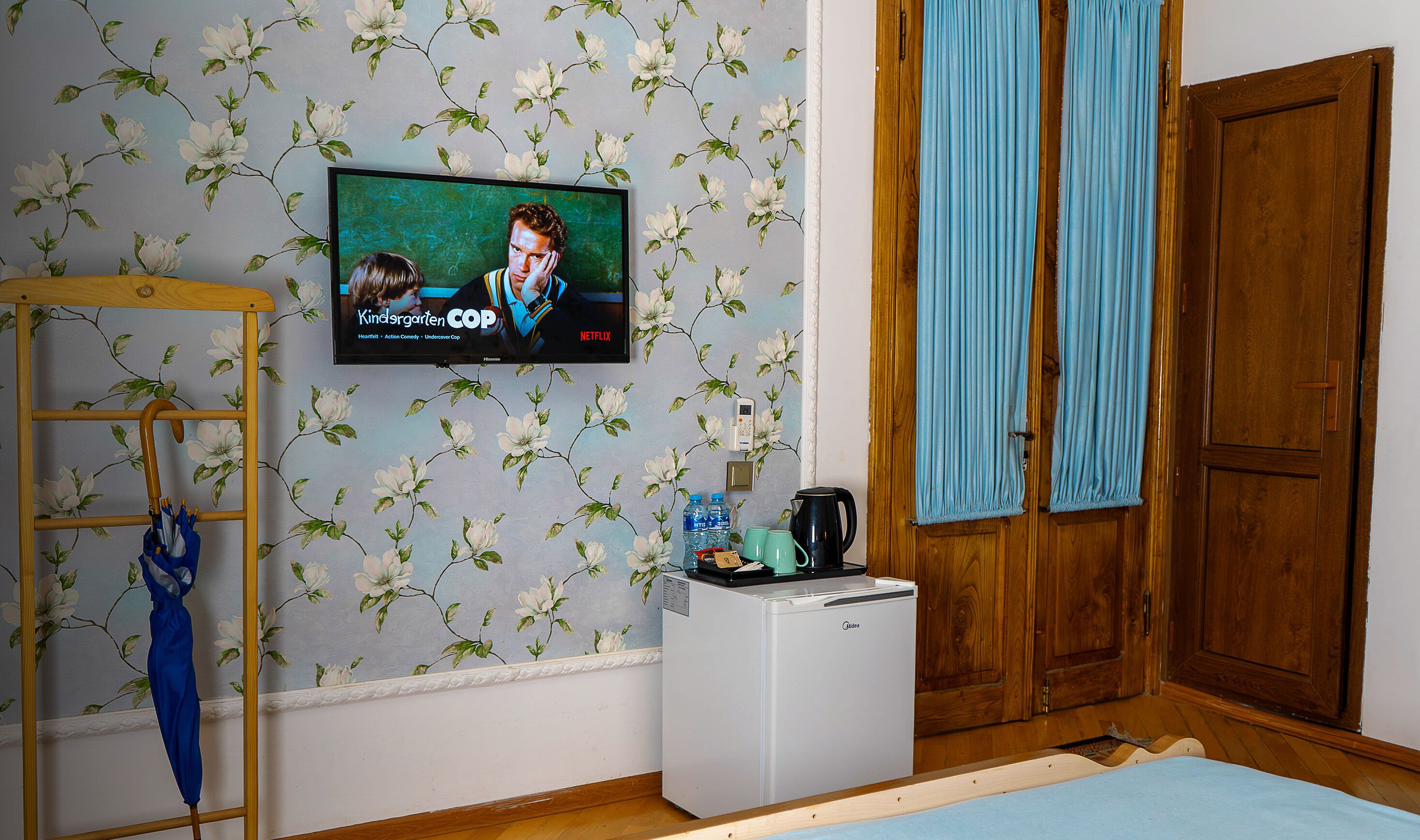 Quarto com papel de parede floral, uma televisão exibindo 'Kindergarten COP', uma pequena geladeira e uma porta de madeira.