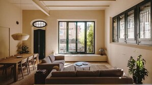 A modern living room with a sofa, coffee table, and dining area. There is a large window with a view of buildings and greenery outside. - Antigua Fábrica Descals, Edificio Histórico (Olot)