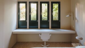 A bedroom with a large bed, a desk with a chair, and three windows with black frames. - Antigua Fábrica Descals, Edificio Histórico (Olot)