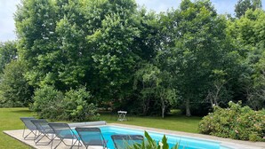 Une piscine avec des chaises longues et une pelouse entourée d’arbres.