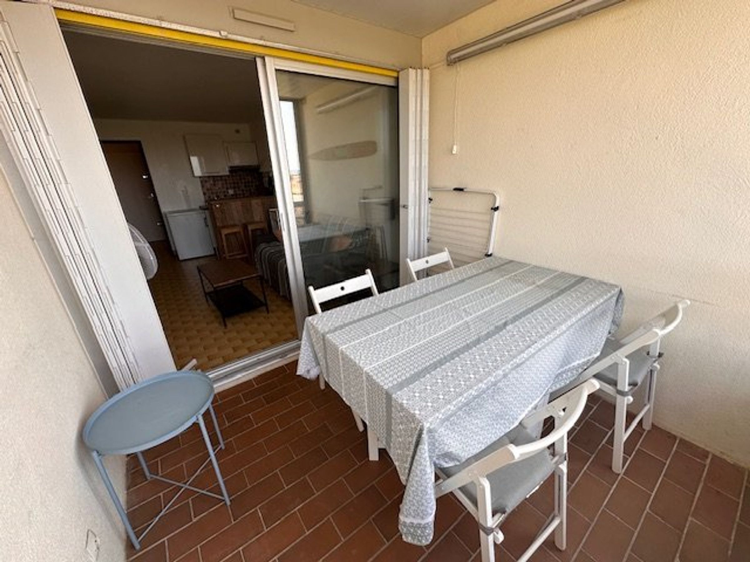 Un petit balcon avec une table et des chaises, une porte coulissante donnant sur un espace intérieur comprenant une cuisine, et une rambarde blanche.