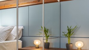 A modern bedroom with a skylight, a bed with white bedding, a bedside table with two potted plants, and a mirror reflecting the room.