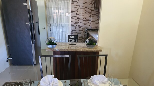 Uma área de refeições com uma mesa posta para dois, dois copos de vinho, um pequeno vaso de planta e uma placa com a inscrição 'WINE O'CLOCK'.