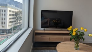 A modern room with a large window, a flat-screen TV on a wooden stand, a vase with yellow flowers, and a view of a building outside. - Trendy Studio at the Barracks on Bree (Cape Town)