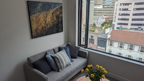 A modern living room with a grey sofa, a painting, and a view of city buildings through a large window. - Trendy Studio at the Barracks on Bree (Cape Town)