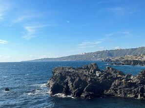 A coastal view with rocky formations and a distant hillside with buildings. - Rifugio di Stazzo Holiday Home for Groups of 20 People (Acireale)