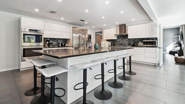 Una cocina moderna con armarios blancos, una isla central con taburetes de bar, electrodomésticos de acero inoxidable y un salpicadero oscuro.
