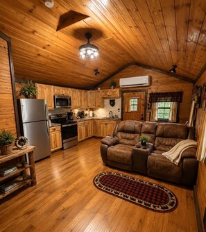 Un intérieur de chalet chaleureux avec un plafond en bois, un coin cuisine doté d’appareils modernes, un canapé en cuir et une cheminée.