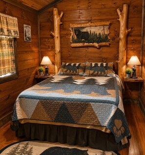 Ein Schlafzimmer in einer Blockhütte mit einem Bett, zwei Nachttischen, einem Wandbild und einem Fenster mit Vorhängen.