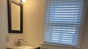 Een badkamer met toilet, wastafel en spiegel. Er is een raam met zonwering en een lichtschakelaar aan de muur.