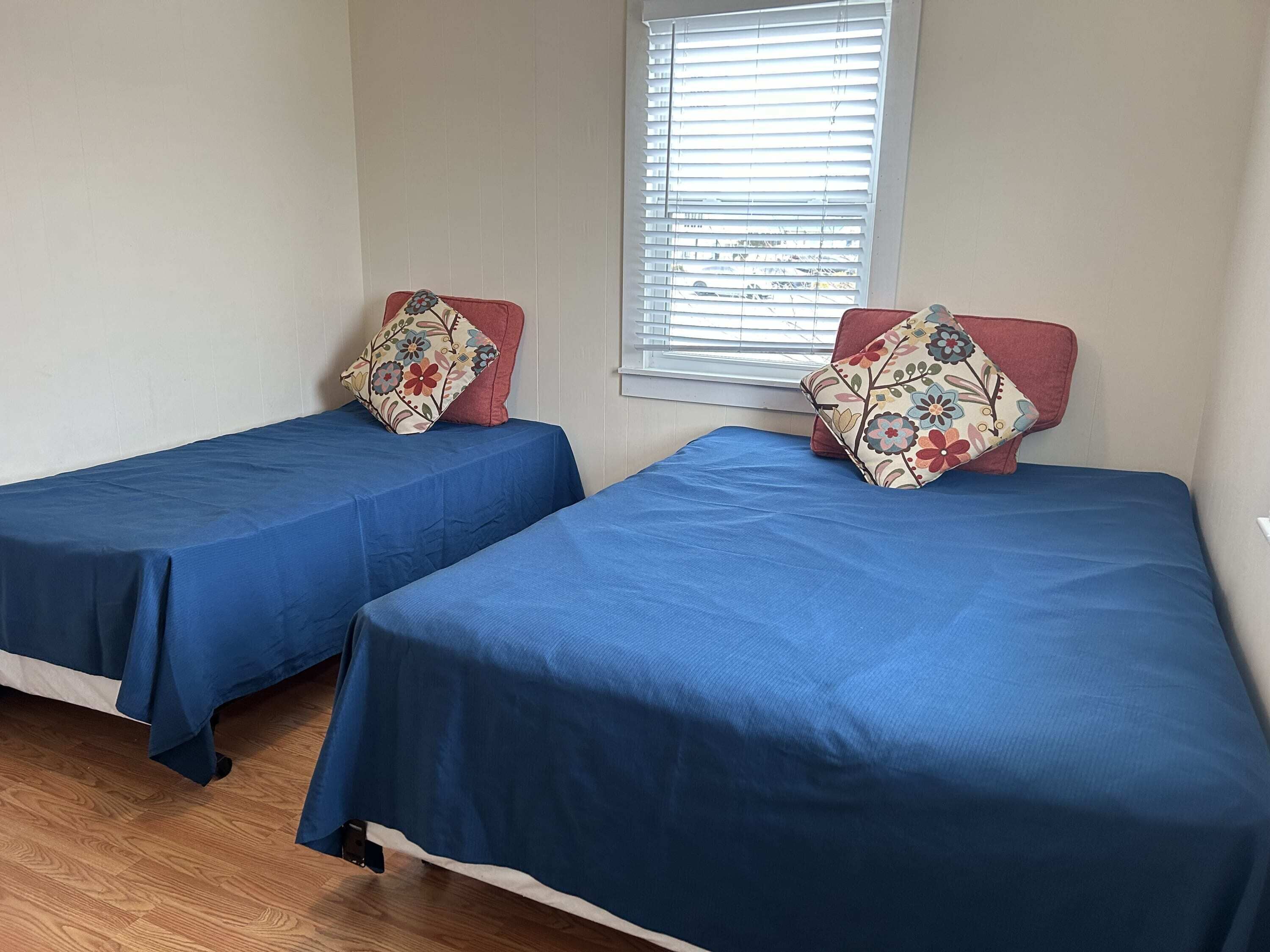 Twee enkele bedden met blauw beddengoed en kussens met een bloemenprint, een raam met zonwering en een houten vloer.