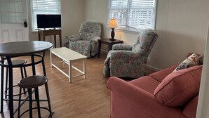 Un salon avec un plancher en bois, une table ronde entourée de tabourets, un canapé, un fauteuil, un téléviseur posé sur un meuble et une lampe sur une table d’appoint.