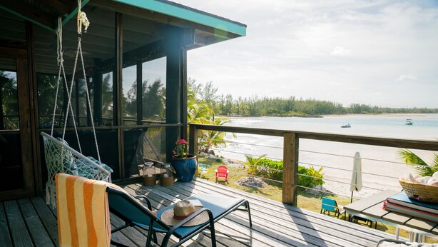 Una terraza de madera con una silla colgante, una silla de descanso y vistas a la playa y barcos.