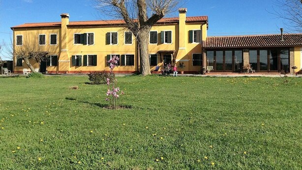Un edificio amarillo con techo rojo, rodeado de un césped con flores y un árbol.