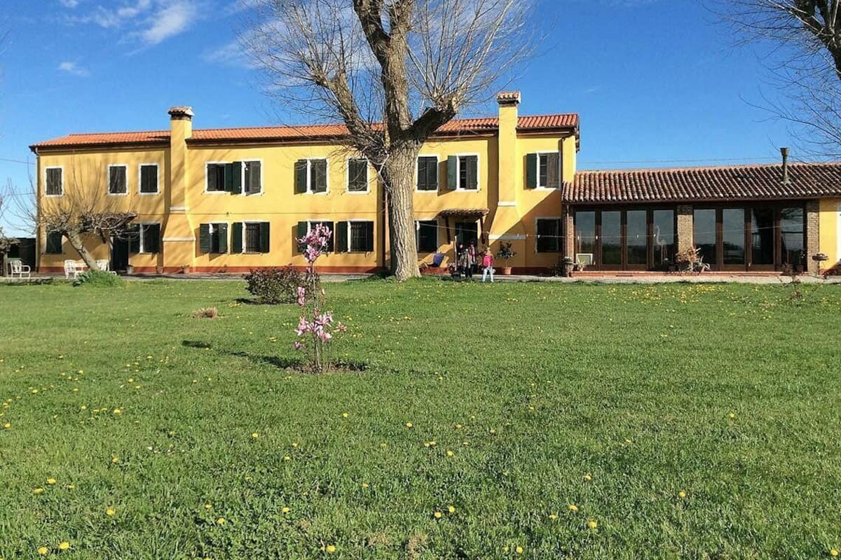 A yellow building with a red roof, surrounded by a grassy lawn with flowers and a tree.