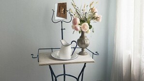 Una habitación con un cuadro enmarcado que muestra una escena de canal, una pequeña mesa con un jarrón de flores, una jarra blanca y un marco de fotos.