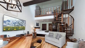 A spacious living area with a curved staircase, a large flat-screen TV, a wooden coffee table, and a sofa with patterned cushions. - The Alpine Line | AC, Pool, Hot Tub, Golf & Ski (Durango)