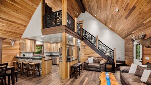 Amplia sala de estar con techo alto, paredes de madera, cocina equipada con electrodomésticos modernos y una escalera con barandillas decorativas.