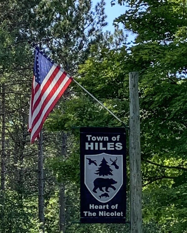 A sign for the Town of Hiles with a bear emblem, an American flag, and trees in the background.