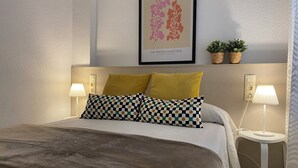 A neatly made bed with a checkered pillow, two pillows in mustard yellow, and a framed artwork above. - Les Doyes 116 (Peníscola)