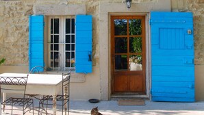 Un bâtiment en pierre avec une porte en bois et des volets bleus, une table avec des chaises en métal, et un chat assis par terre.