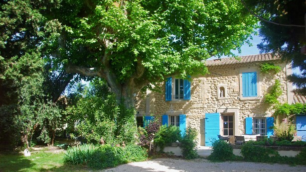 Une maison en pierre avec des volets bleus, entourée d’une végétation luxuriante et bordée d’un chemin de gravier.
