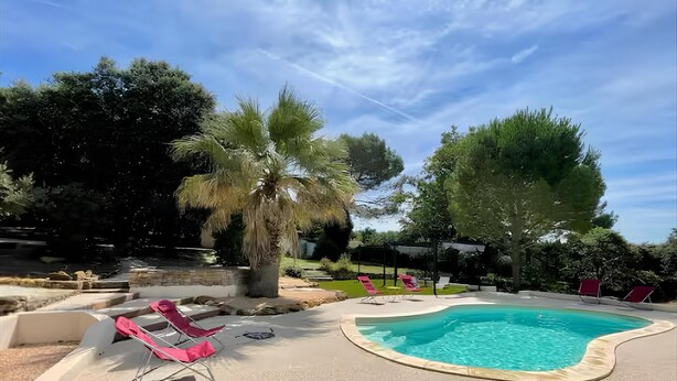 A swimming pool with a curved edge, surrounded by a concrete deck. There are red lounge chairs and a pergola with a white canopy. - Maison Familiale Avec Piscine et Magnifique Jardin, Sud de la France (Piolenc)