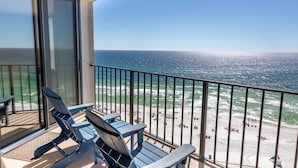 Ein Balkon mit zwei blauen Adirondack-Stühlen, einer Glastür und Blick auf Strand und Meer.