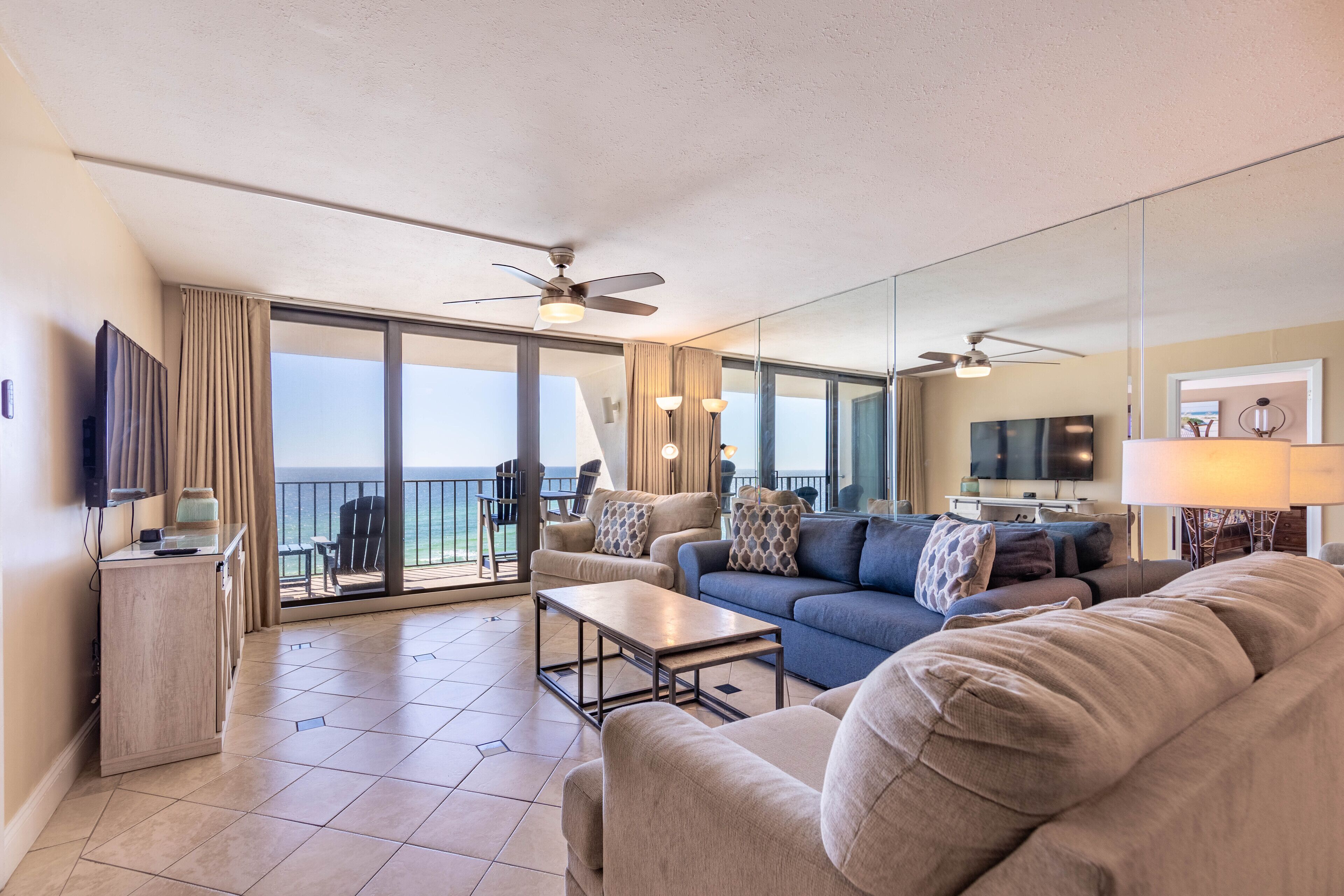 A spacious living area with a sofa, coffee table, and a television. There is a balcony with a view of the ocean.