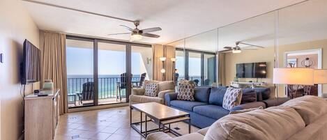 A spacious living area with a sofa, coffee table, and a television. There is a balcony with a view of the ocean.