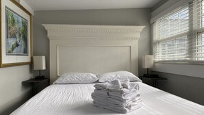 Un lit bien fait, avec du linge de lit blanc et des oreillers, une pile de serviettes pliées, un tableau encadré au mur et une fenêtre avec des stores.