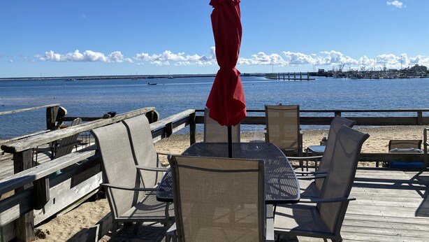 Une terrasse en bois, avec des chaises et une table, donnant sur une mer calme où l’on aperçoit des bateaux et, au loin, une jetée.